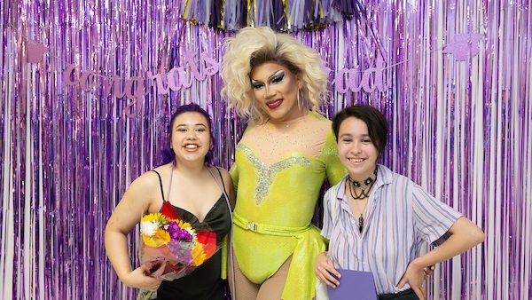 Photo of a lavender grad photo booth with two students posing with a drag artist and flowers. 
