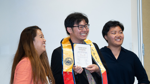 Photo with a male student posing with their APIDA stole and certificate smiling with two APIDA Center staff members